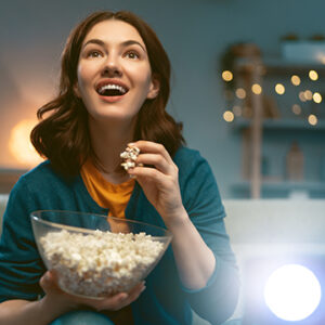 Young woman watching projector, TV, movies with popcorn in the evening. Girl spending time at home.