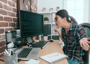Person sitting at a desk in front of two screens displaying computer code, with a laptop, notebook and work accessories, in a brick-walled office space.